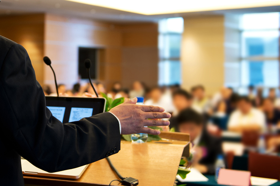 Business man is making a speech in front of a big audience at a conference hall.